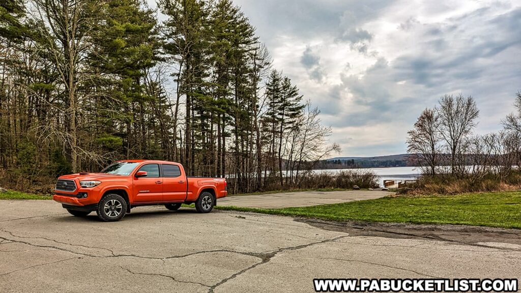 A vibrant orange pickup truck is parked on an asphalt lot, surrounded by towering green trees. In the background, a serene lake stretches into the distance, hinting at the natural beauty of Shohola Falls in Pike County, Pennsylvania. The sky above is a dynamic canvas with patches of blue peeking through the predominant gray-white clouds, suggesting a cool, overcast day. The scene is peaceful, with the truck standing out against the natural palette of the landscape.