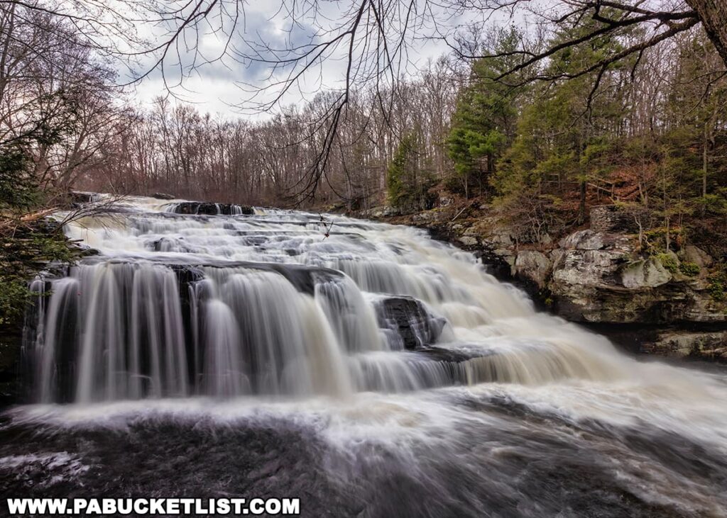 A cascading waterfall at Shohola Falls in Pike County, Pennsylvania, flows vigorously over tiered rocky ledges. The rushing water creates a smooth, silky texture, indicative of a long exposure photograph. Surrounding the falls are bare deciduous trees and evergreens, suggesting a season of early spring. The sky above is a soft mix of white and blue, possibly indicating an overcast day. The natural setting evokes a sense of tranquility and the raw power of nature.