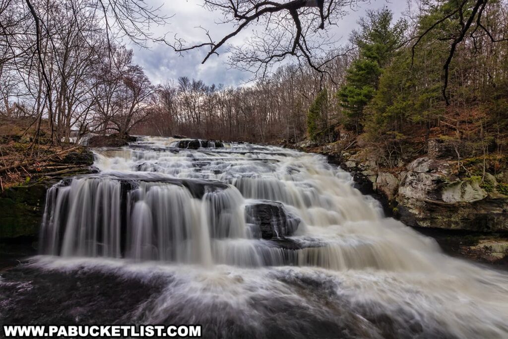 A wide-angle view captures the majestic Shohola Falls in Pike County, Pennsylvania, with water powerfully cascading over a series of natural rock terraces. The falls are nestled in a woodland setting, with leafless trees and evergreens framing the scene, suggesting the image was taken in the cooler months. The water's smooth, white veils are the result of a slow shutter speed, emphasizing the motion and the waterfall's grace. Overcast skies with a hint of blue add a serene mood to this natural landscape.