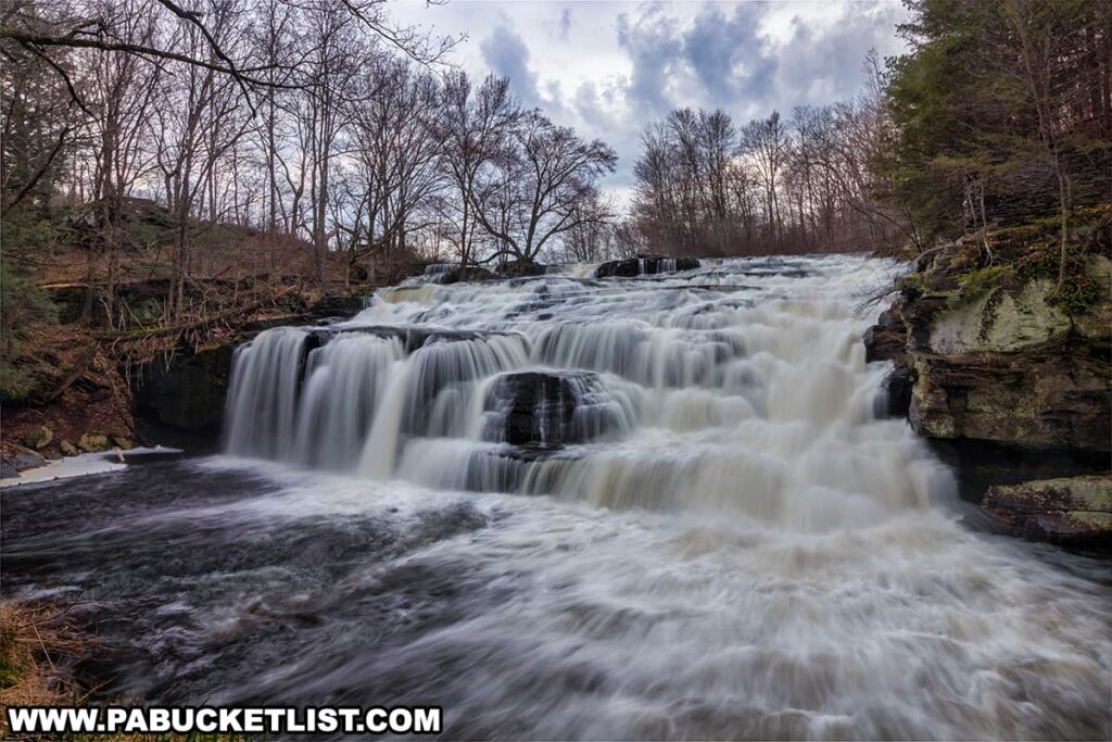 The image showcases the dynamic Shohola Falls in Pike County, Pennsylvania, where the water forcefully tumbles over a multitude of rock ledges. The perspective captures the breadth and power of the falls, framed by the rugged beauty of the surrounding rock faces and a variety of trees, some with bare branches suggesting an early spring or late fall season. Clouds scatter across the sky, casting a soft, diffused light that enhances the waterfall's white frothy cascades. The scene conveys a natural, untouched wilderness.