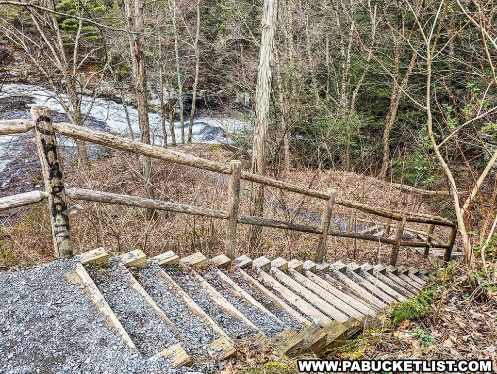 The image captures a rustic wooden staircase leading down to a viewing area of Shohola Falls in Pike County, Pennsylvania. The stairs are bordered by a simple wooden fence, both showing signs of natural weathering. Leafless trees are scattered throughout the area, suggesting that the season is either fall or winter. In the background, glimpses of the waterfall can be seen through the trees, with the water appearing white and frothy as it rushes over rocks. The overcast sky implies a cool and possibly damp day, adding to the earthy and natural ambiance of the scene.