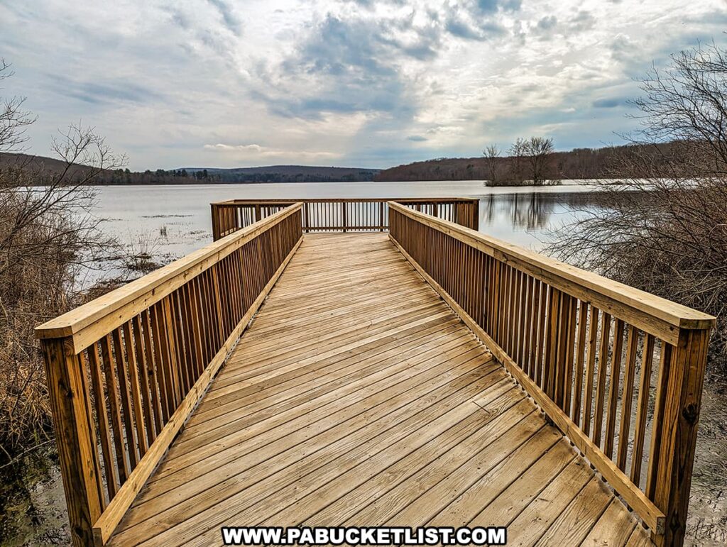 The image depicts a well-constructed wooden walkway leading to an observation deck overlooking a lake at Shohola Falls in Pike County, Pennsylvania. The deck provides an unobstructed view of the tranquil water and a partially submerged cluster of trees, reflecting the calm, scenic environment. The sky above is a dramatic expanse of white and gray clouds, suggesting an overcast day. The surrounding landscape is in a state of winter dormancy with bare trees, and the wooden structure offers a sturdy vantage point for visitors to take in the serene beauty of the lake.