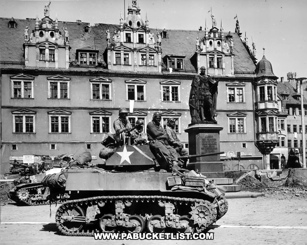 A black and white historical photograph depicting an M5A1 Stuart tank with soldiers aboard, parked in a European square with ornate buildings in the background, alongside a statue of a historic figure on a pedestal.