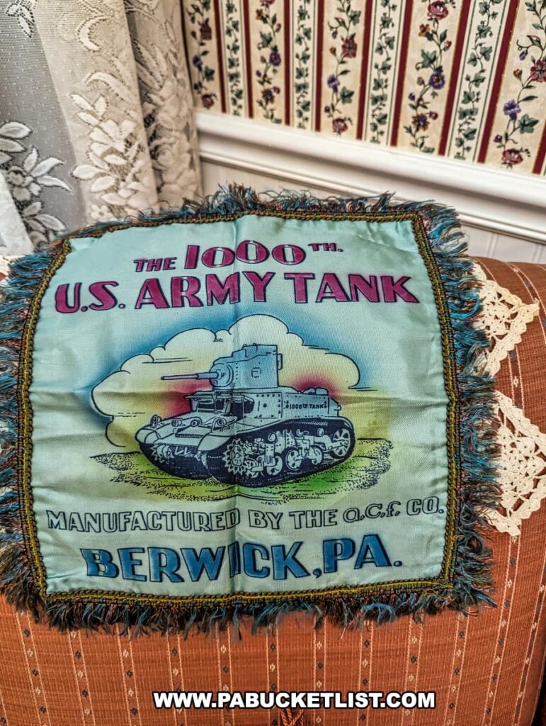 A commemorative pillow at the Stuart Tank Museum in Berwick, Pennsylvania, features colorful embroidery celebrating the 1000th U.S. Army tank manufactured by the American Car and Foundry Company in Berwick, PA, displayed against a traditional fabric chair with floral wallpaper in the background.
