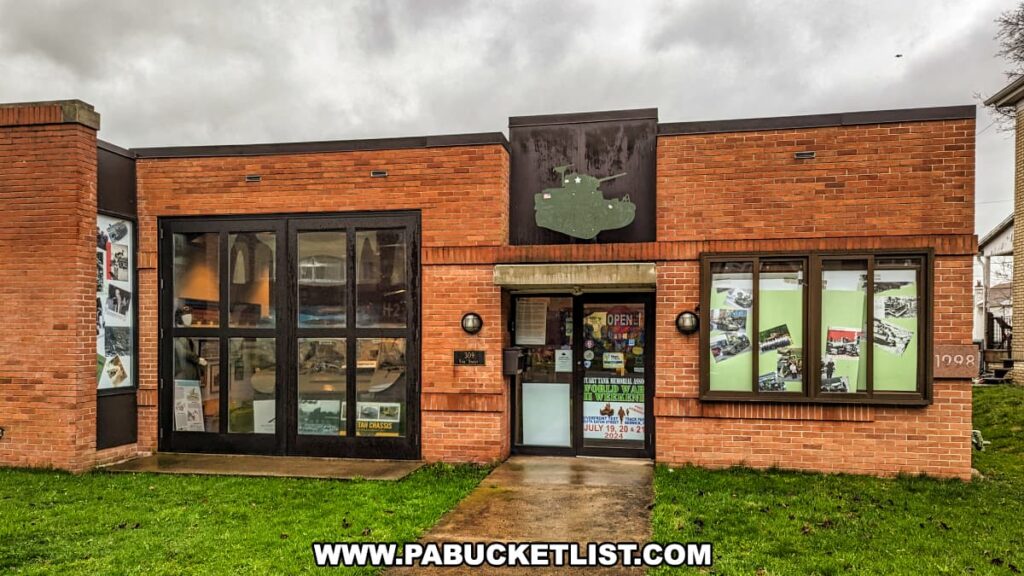 The front view of the Stuart Tank Museum in Berwick, Pennsylvania, showcasing its brick facade with a tank silhouette sign above the entrance, large glass windows with visible displays inside, and an "OPEN" sign inviting visitors to explore the history within.