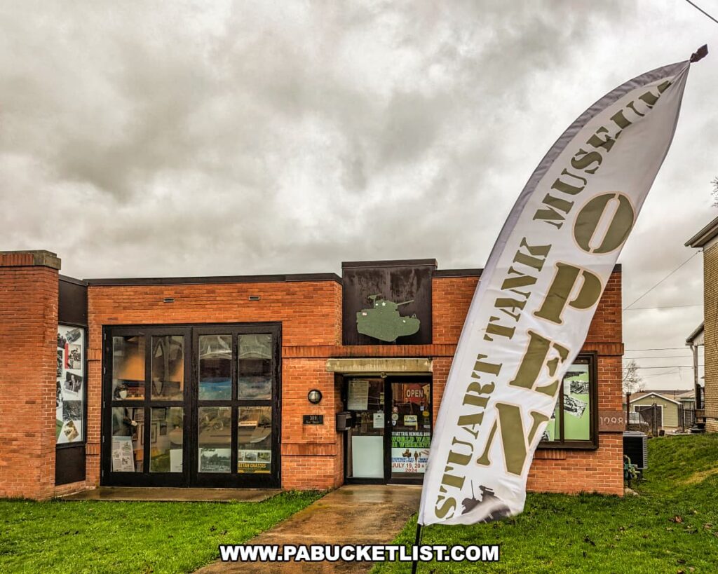 The entrance to the Stuart Tank Museum in Berwick, Pennsylvania, is marked by a large vertical 'OPEN' banner next to the building's brick facade, with the silhouette of a Stuart tank displayed prominently above the front doors under an overcast sky.