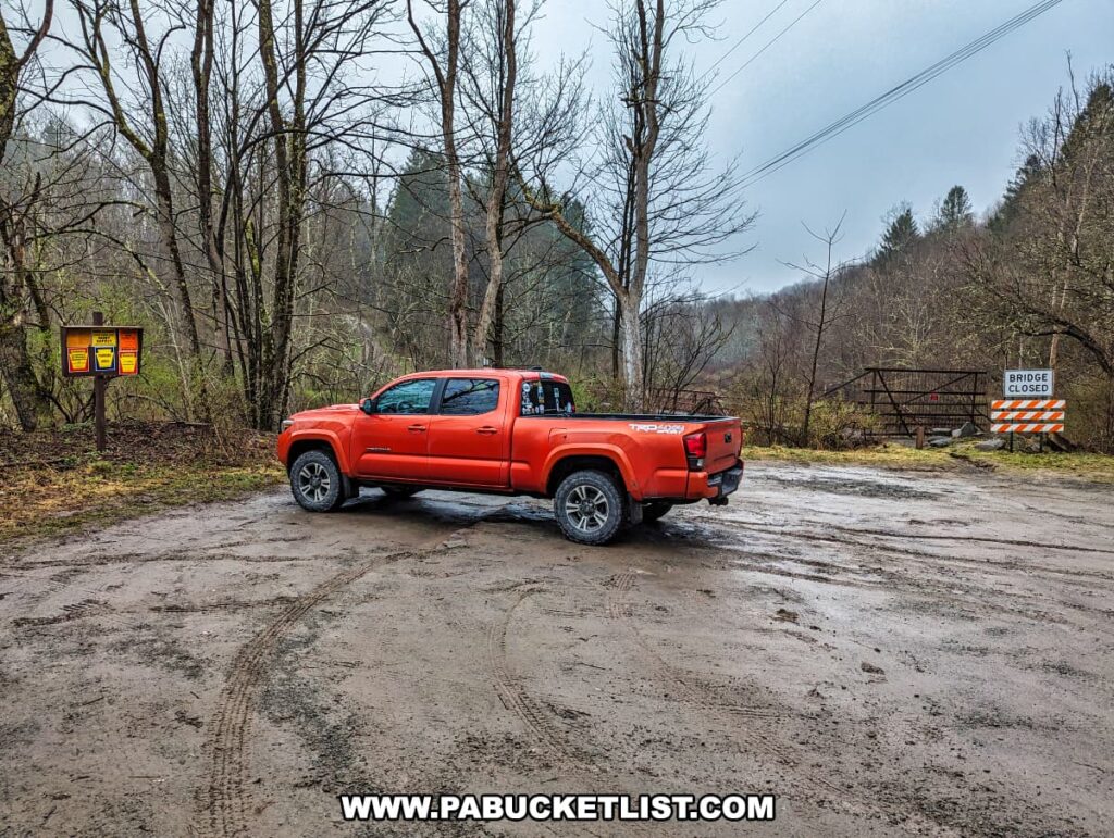 A vibrant orange pickup truck is parked on a muddy gravel lot near Tanners Falls on State Game Lands 159 in Wayne County, Pennsylvania. The truck faces a bridge marked with a "BRIDGE CLOSED" barrier. Leafless trees and overcast skies indicate a barren, possibly cold day. In the background, hints of a forested area suggest the proximity to natural attractions, such as the waterfall and historical tannery ruins dating back to 1931.