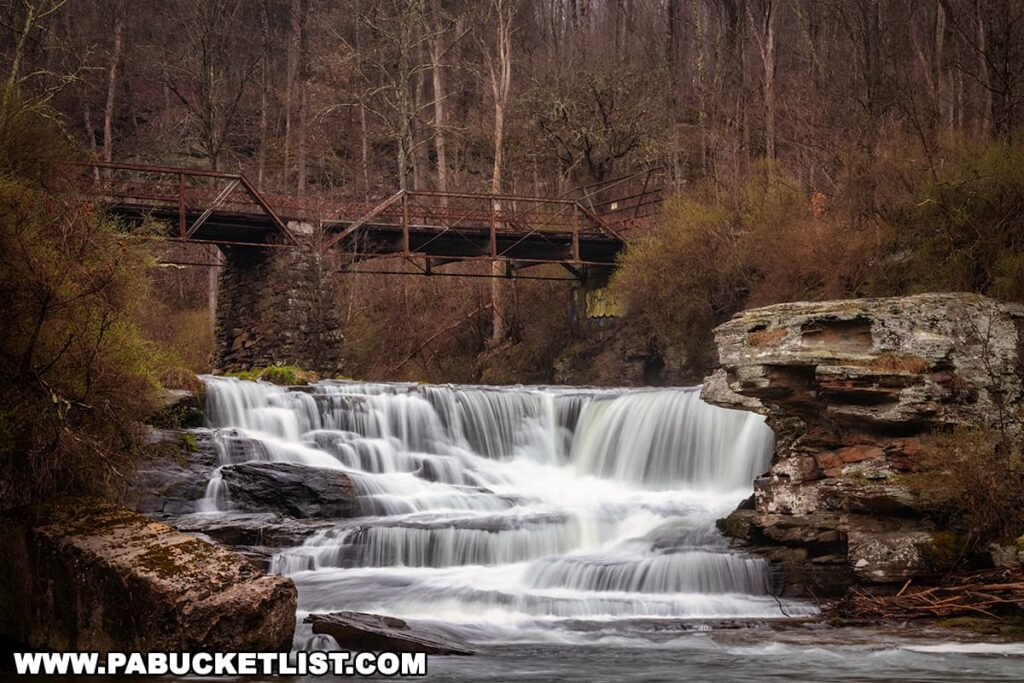 The serene Tanners Falls in Wayne County, Pennsylvania flows gracefully over stratified rock ledges, surrounded by the awakening spring landscape of State Game Lands 159. A rustic metal bridge, showcasing utilitarian architecture, spans across the stream in the background, creating a picturesque scene that juxtaposes the man-made structure with the waterfall's natural beauty. The waterfall, part of the historic site where a tannery once operated until 1931, is enveloped by trees that are just beginning to bud, hinting at the rebirth of the forest in early spring.