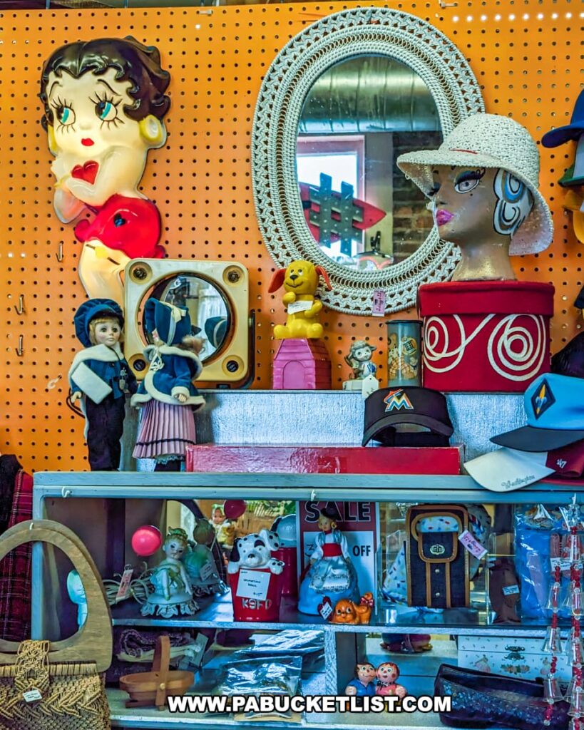 A vibrant display at the Burning Bridge Antiques Market in Lancaster County, PA, featuring a variety of vintage and collectible items. Prominently displayed is a large Betty Boop light-up figure on an orange pegboard background. The display includes an eclectic mix of items such as a mannequin head with a hat, a decorative oval mirror with a crocheted frame, vintage dolls, toy figures, and assorted hats. Shelves below hold more collectibles including ceramic figurines, a wicker purse, and various trinkets, creating a colorful and nostalgic atmosphere within this extensive antique store.