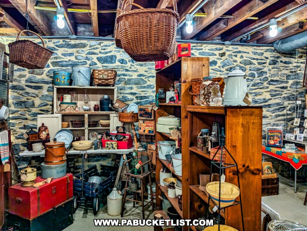 A cozy, rustic basement area at the Burning Bridge Antiques Market in Lancaster County, PA. The space features a variety of vintage household items and kitchenware, including enamelware, ceramic bowls, tin containers, and woven baskets. Wooden shelves and a stone wall backdrop add to the nostalgic feel of the setting. Hanging baskets, old-fashioned lanterns, and colorful tins are displayed creatively, showcasing the diverse selection of antiques available from the many vendors in this expansive store. Exposed beams and industrial lighting give the space a warm, inviting atmosphere.