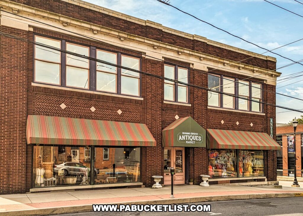 The exterior view of the Burning Bridge Antiques Market in Lancaster County, PA, showcases a large, brick building with two stories of windows. The entrance features striped awnings above large display windows, giving a glimpse of the antiques inside. The sign above the entrance reads "Burning Bridge Antiques Market," indicating the store's name. The building's brickwork includes decorative patterns, and the sidewalk in front has street parking available. This expansive antique store houses over 200 vendors and is a prominent feature in the Columbia area.