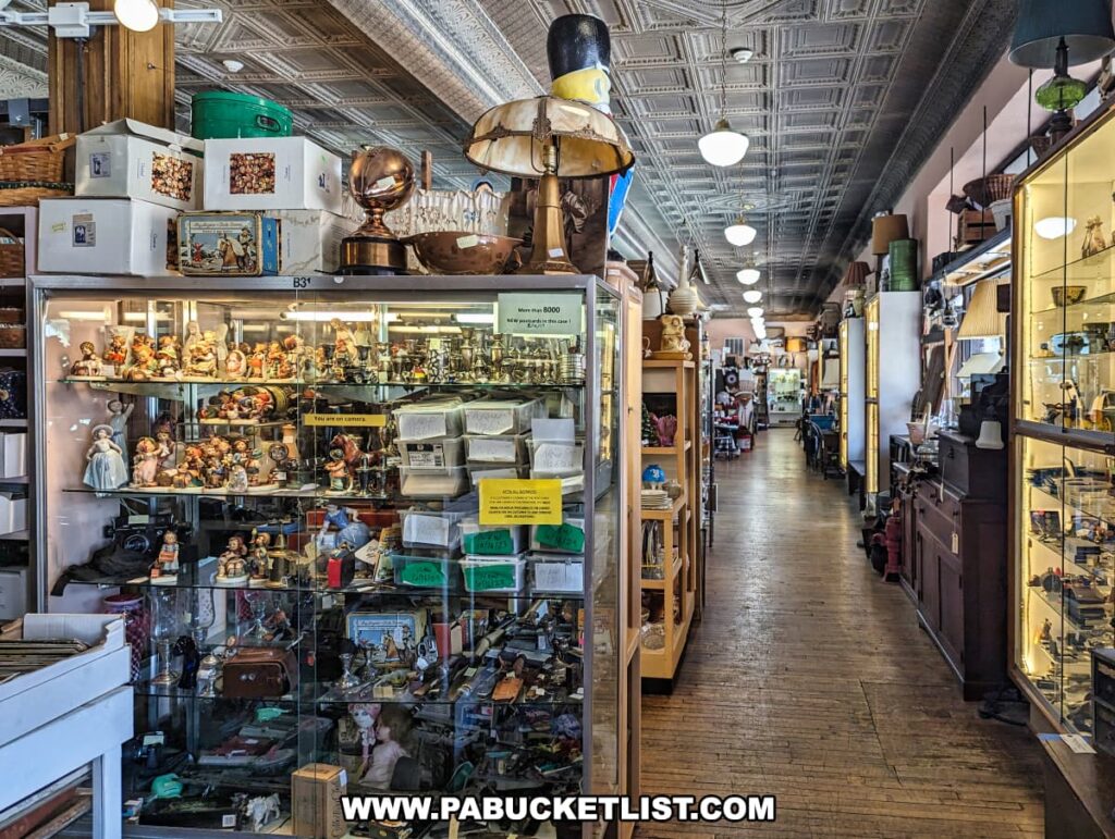 A long aisle inside the Burning Bridge Antiques Market in Lancaster County, PA, showcasing a variety of antique items displayed in glass cases and on wooden shelves. The display cases are filled with vintage figurines, collectibles, and other small treasures, while the shelves hold larger items, including lamps and decorative pieces. The store features a high, ornate ceiling with hanging lights and a wooden floor, contributing to its historic charm. The aisle stretches into the distance, lined with more antiques and collectibles, reflecting the store's vast collection from over 200 vendors.