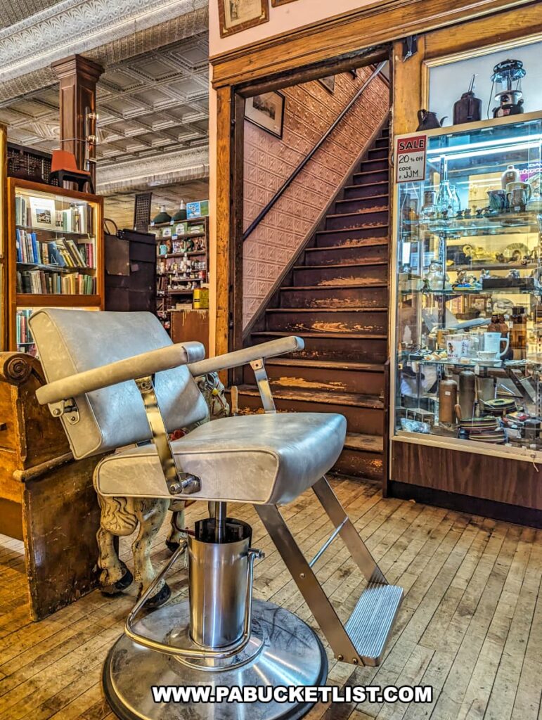A vintage barber chair is prominently displayed near a wooden staircase at the Burning Bridge Antiques Market in Lancaster County, PA. The chair features a metallic frame and a padded seat and backrest. Behind it, an old wooden staircase with worn steps leads to the upper floor. To the right, a glass display case filled with various antique items, including jewelry and small collectibles, is visible. The store's interior showcases a tin ceiling, wooden floors, and bookshelves, adding to the nostalgic and historic charm of this extensive antique market with over 200 vendors.