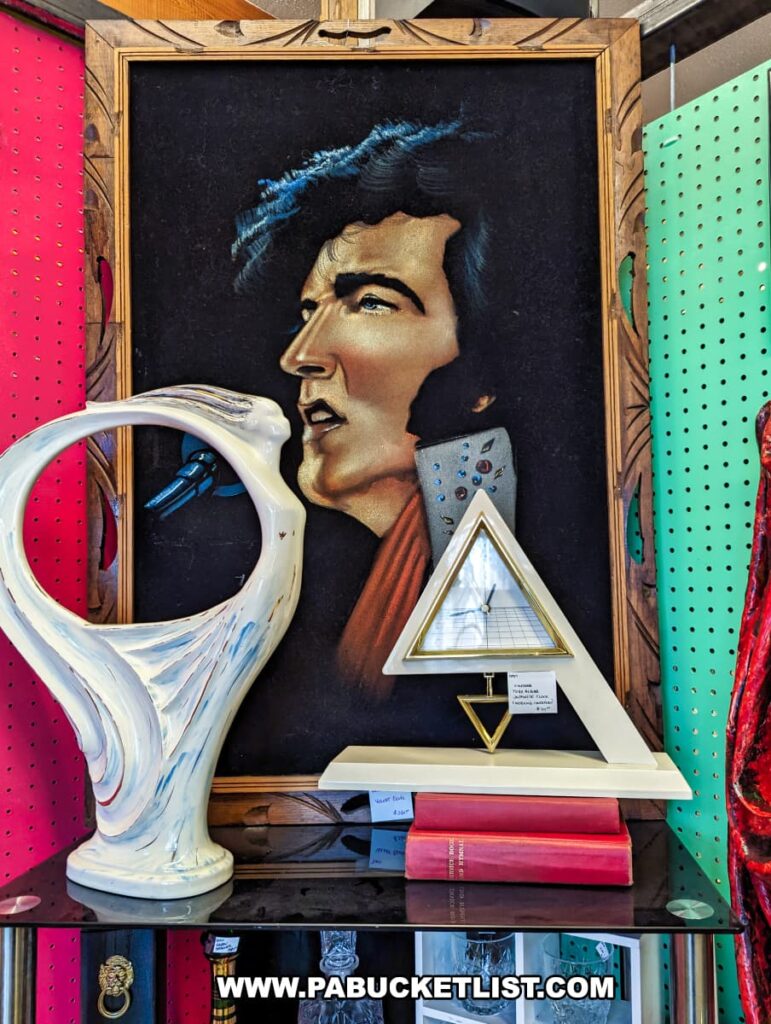 A display at the Burning Bridge Antiques Market in Lancaster County, PA, featuring a framed velvet painting of Elvis Presley in a detailed wooden frame. The painting depicts Elvis in a side profile, singing into a microphone, adorned in a studded outfit. In front of the painting are various decorative items, including a white ceramic abstract sculpture and a modern triangular clock with a gold stand. Red books and other small collectibles are also part of the display, all set against a vibrant background of red and green pegboards, contributing to the eclectic and nostalgic atmosphere of the antique market.