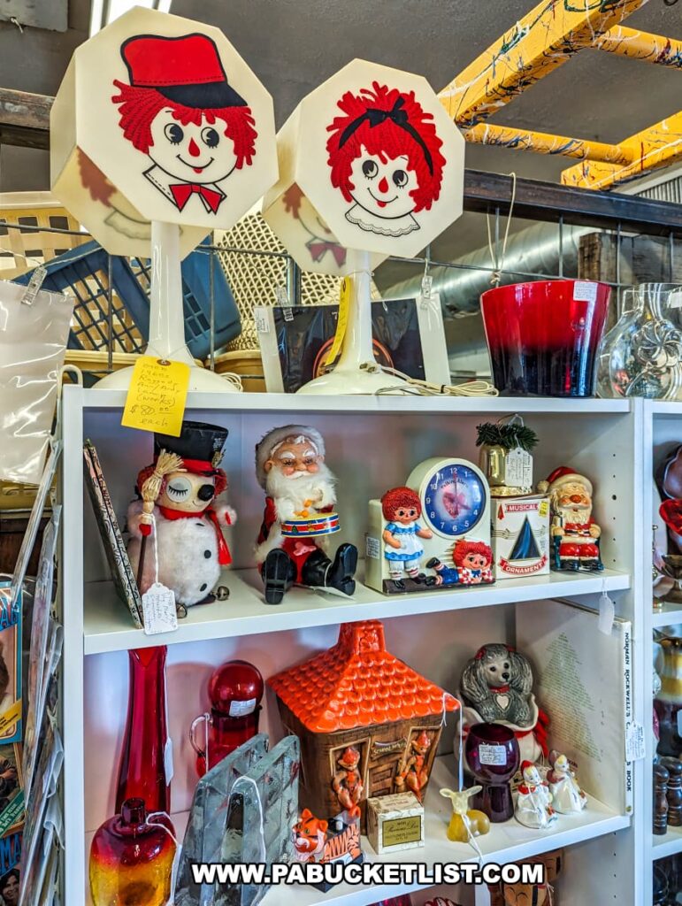 A colorful display of vintage items at the Burning Bridge Antiques Market in Lancaster County, PA. The top shelf features two lamps with shades depicting Raggedy Ann and Andy faces. Below, shelves are filled with various nostalgic items, including a plush Santa Claus, a musical clock with a Christmas scene, a ceramic gingerbread house, vintage glassware, and a variety of figurines and ornaments. The eclectic collection showcases the wide range of collectibles available from the numerous vendors at this extensive antique market. The vibrant and whimsical items create a charming and nostalgic atmosphere.