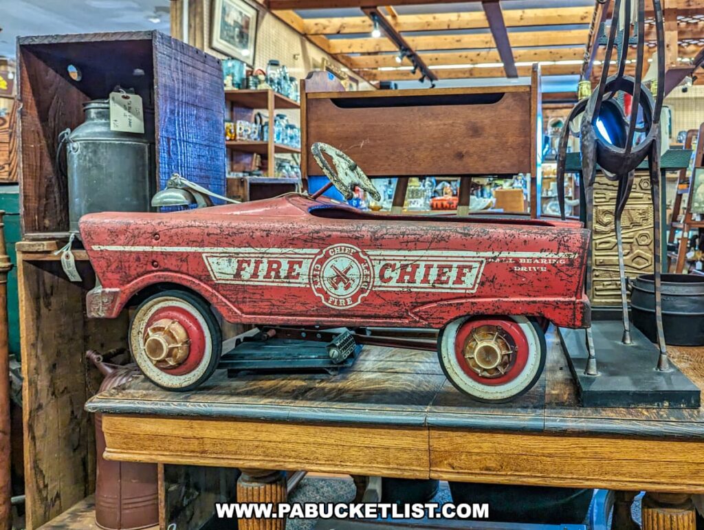 A vintage red pedal car labeled "Fire Chief" on display at the Burning Bridge Antiques Market in Lancaster County, PA. The car, showing signs of age with worn paint and rust spots, adds to its nostalgic charm. It features a detailed design with the words "Fire Chief" and "Ball Bearing Drive" prominently displayed on the side. The car is positioned on a wooden table among other antique items, including a metal milk can and various wooden furniture pieces, highlighting the diverse collection available from the numerous vendors at this expansive antique market.