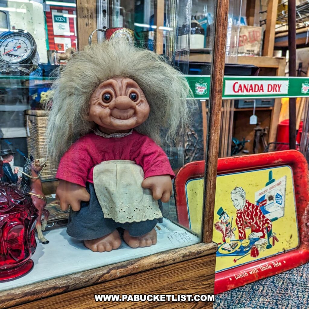 A vintage troll doll on display at the Burning Bridge Antiques Market in Lancaster County, PA. The troll doll features wild, white hair and is dressed in a red shirt with a white apron and blue pants. It is displayed inside a glass cabinet along with other vintage items, including a red glass bottle and a toy horse. Next to the cabinet is a colorful metal tray with a "Canada Dry" logo and an illustration of a man having lunch with a clown. The display highlights the variety of quirky and nostalgic collectibles available from the numerous vendors at this expansive antique market.