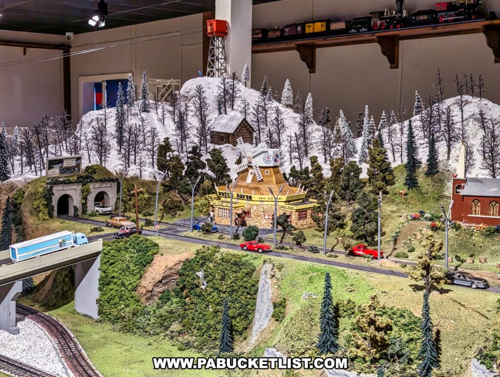 A detailed model train display at the Choo Choo Barn in Lancaster County, PA, showcasing a miniature replica of the Dutch Haven Shoo-Fly Pie Shop surrounded by lush greenery and a snow-capped mountain. The scene includes multiple animated figures, vehicles on the road, and a train track running through a tunnel beneath a bridge. The intricate details and realistic features capture the charm and essence of the local landmarks in the miniature world.