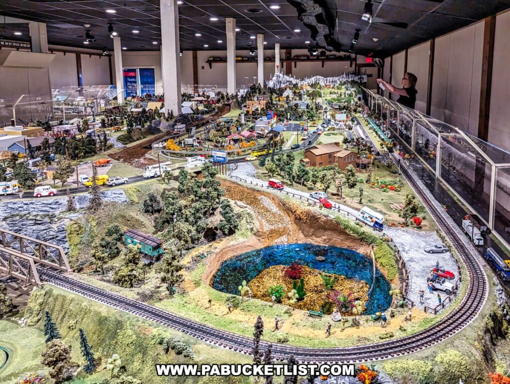 An expansive model train display at the Choo Choo Barn in Lancaster County, PA, featuring a detailed landscape with a miniature lake, surrounded by lush greenery and various buildings. The scene includes multiple train tracks, roads with vehicles, and animated figures engaged in different activities. The intricate display stretches out in a wide view, capturing the diverse and dynamic elements of the miniature world.