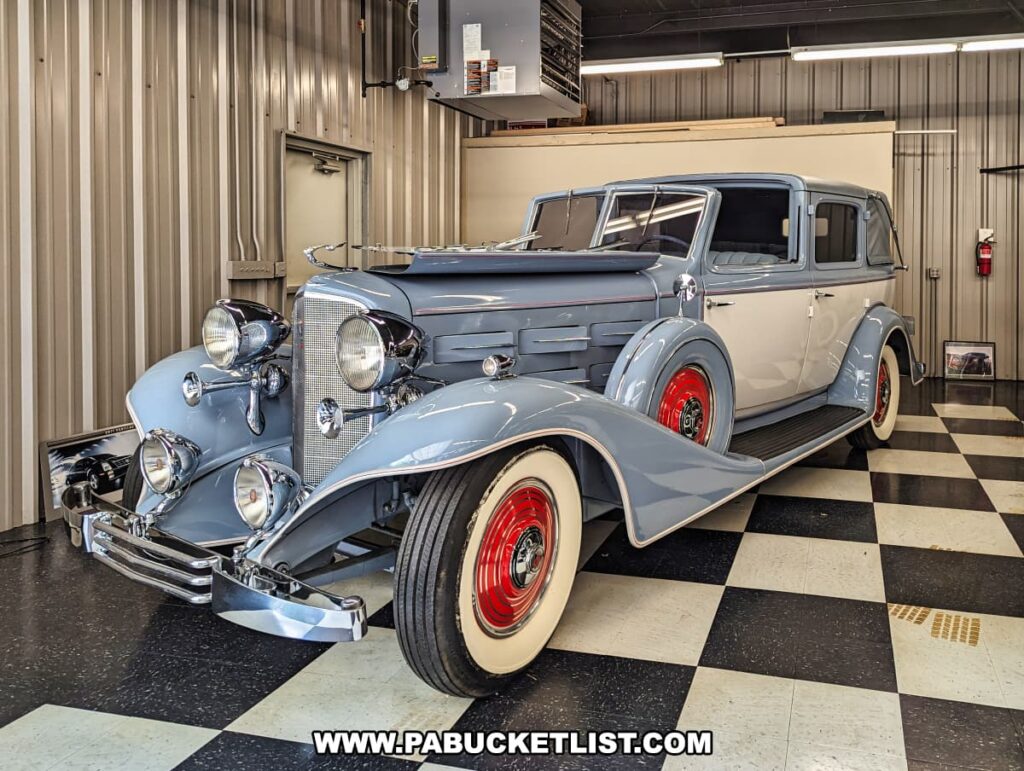 A pristine 1933 Cadillac Landaulet model on display at the Greenberg Cadillac Museum in Jefferson County, PA. The car features a stylish two-tone gray and white exterior with striking red wheels. The well-maintained vehicle highlights the museum's dedication to preserving the elegance and historical significance of classic Cadillacs. The checkered floor and industrial setting provide a fitting backdrop for this vintage automobile.