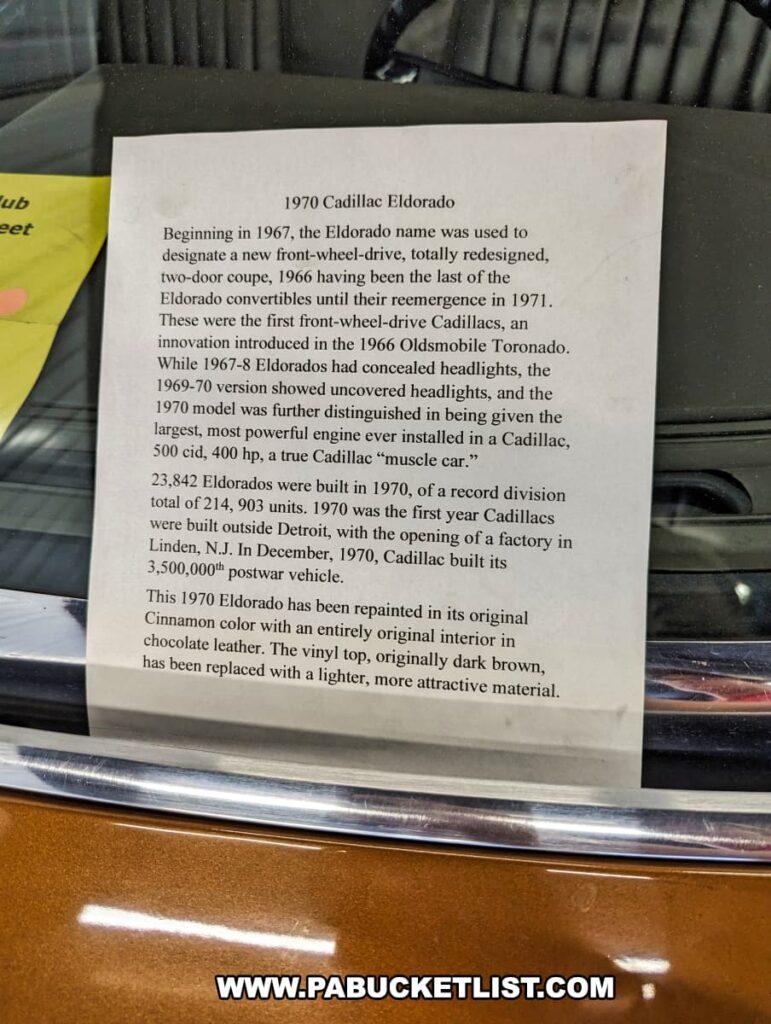 An informational sign detailing the history of the 1970 Cadillac Eldorado on display at the Greenberg Cadillac Museum in Jefferson County, PA. The sign highlights the Eldorado's introduction in 1967 as a front-wheel-drive vehicle, its powerful 500 cubic inch engine, and its unique design features, including uncovered headlights. It also notes that the car has been repainted in its original Cinnamon color with a new, lighter vinyl top and an interior featuring original chocolate leather. The background shows part of the car’s bronze exterior, emphasizing the museum's dedication to preserving the detailed history of each vehicle.
