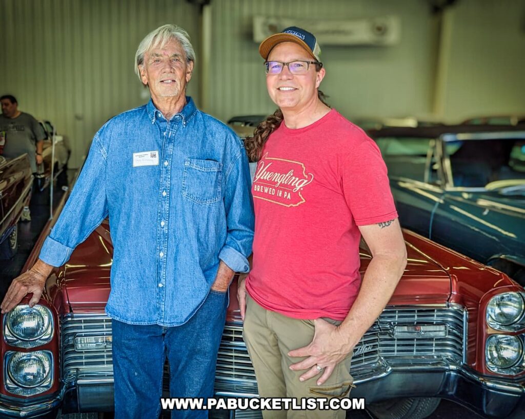 Dr. Steven Greenberg, founder of the Greenberg Cadillac Museum, stands next to Rusty Glessner, the author of PA Bucket List, in front of a classic red Cadillac. The photo is taken inside the museum, showcasing the vintage cars in the background. Dr. Greenberg is dressed in a blue denim shirt, while Rusty Glessner wears a red Yuengling t-shirt and a baseball cap, both smiling for the camera. The image captures a moment of shared appreciation for the extensive Cadillac collection.