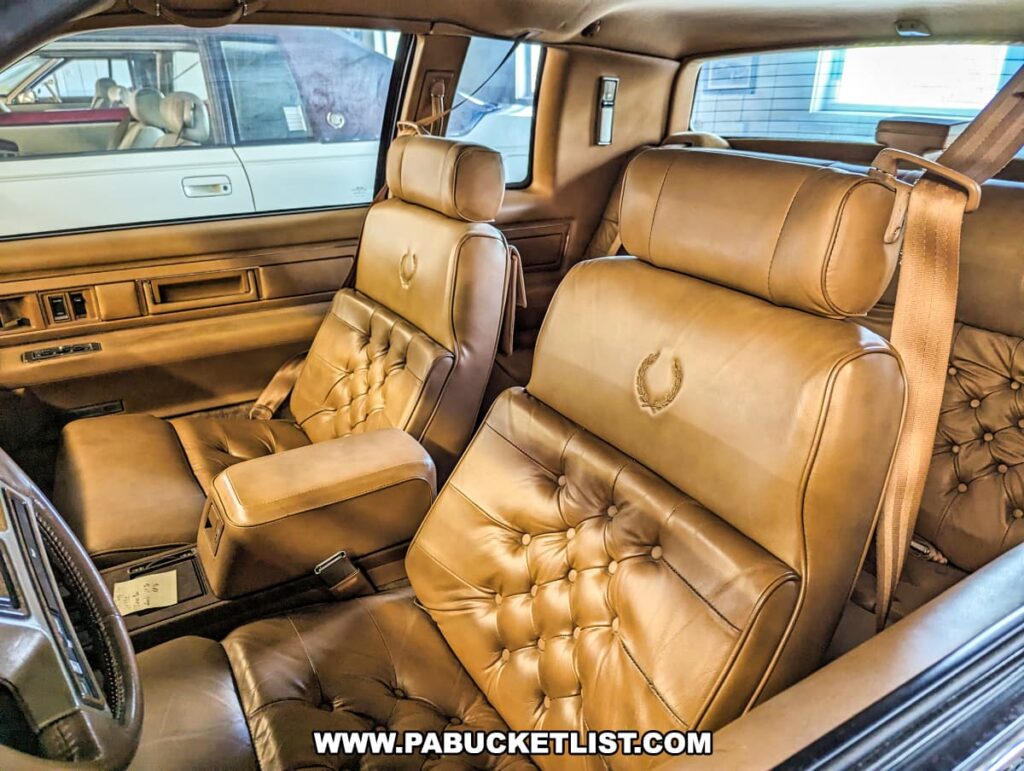 The interior of a modern Cadillac on display at the Greenberg Cadillac Museum in Jefferson County, PA. The car features luxurious tan leather seats with tufted upholstery and embroidered Cadillac crests on the headrests. The interior design emphasizes comfort and elegance, showcasing the attention to detail and craftsmanship characteristic of Cadillac vehicles. Other vintage Cadillacs are visible through the windows, highlighting the museum's extensive collection.