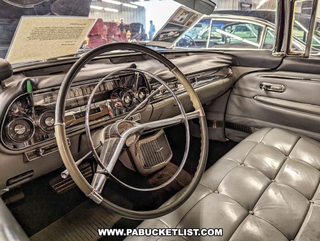 The interior of a vintage Cadillac on display at the Greenberg Cadillac Museum in Jefferson County, PA. The dashboard features classic instrumentation and a large steering wheel with chrome accents. The car's interior is upholstered in luxurious gray leather with tufted seating, reflecting the elegance and craftsmanship of Cadillac vehicles from this era. Informational plaques are visible on the dashboard, providing historical context for visitors. Other vintage cars can be seen in the background, emphasizing the museum's extensive collection.