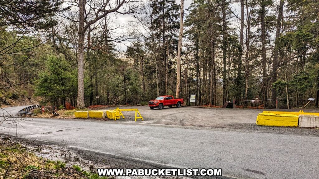 A view of the parking area for Hackers Falls in Pike County, Pennsylvania, located along Raymondskill Road. The area features a small gravel lot with a single red truck parked. Yellow concrete barriers and a sign mark the entrance to the trailhead, surrounded by tall trees and dense forest, creating a secluded and inviting starting point for hikers.