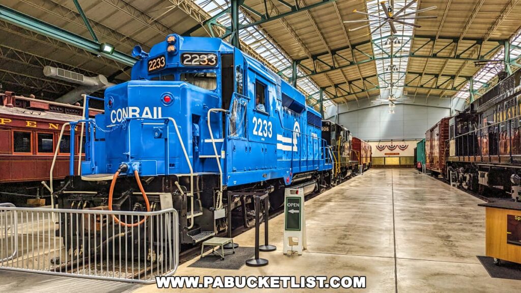 The image captures a vivid blue Conrail locomotive prominently displayed at the Railroad Museum of Pennsylvania in Strasburg, Lancaster County. The locomotive, numbered 2233, is showcased in a large, well-lit exhibit hall alongside a variety of other historic railcars, including a Pennsylvania Railroad passenger car. The scene is set under a high industrial ceiling with skylights, enhancing the natural light that floods the space. The museum’s spacious interior and the meticulous arrangement of the locomotives and railcars highlight the historical significance and engineering marvels of the railroad industry.