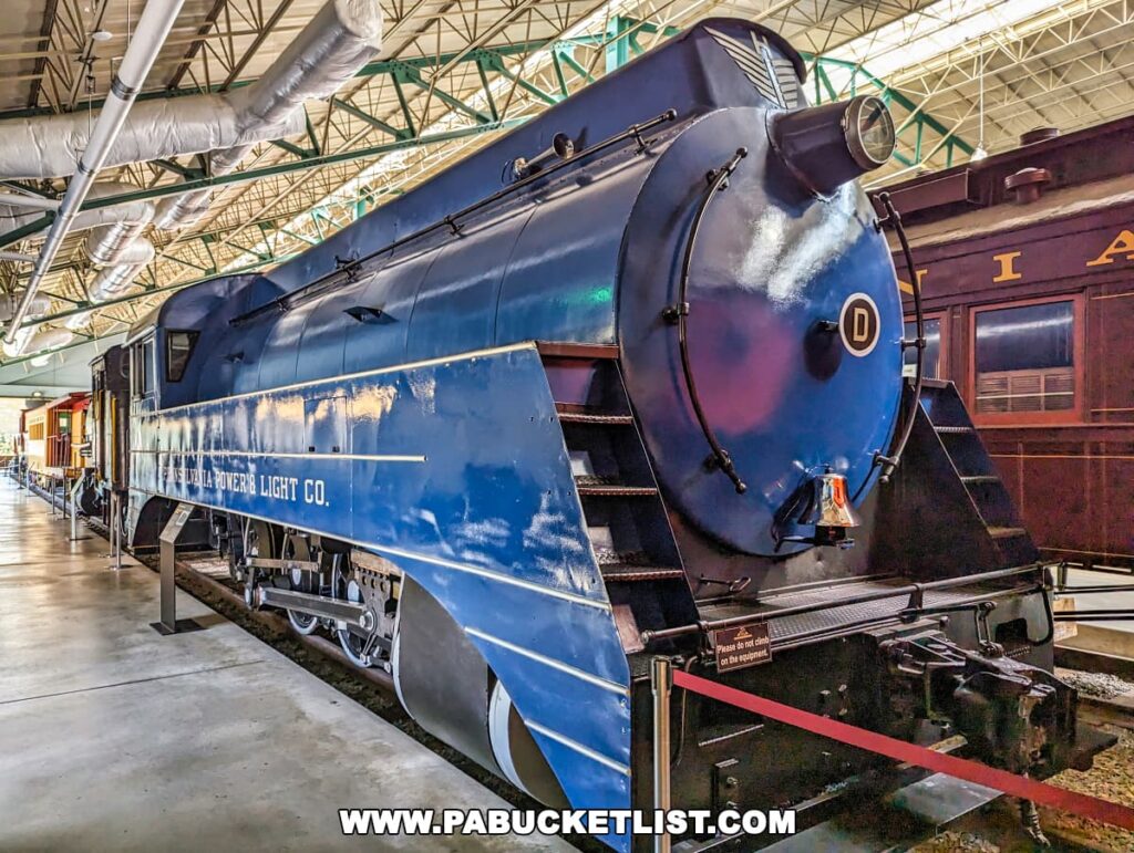 The image showcases a vibrant blue steam locomotive from the Pennsylvania Power & Light Co., exhibited at the Railroad Museum of Pennsylvania in Strasburg, Lancaster County. The locomotive, with its polished, streamlined design and prominent round headlamp, is a striking example of mid-20th-century industrial steam engine technology. Its deep blue color and large white lettering along the side stand out against the indoor setting of the museum, highlighted by the natural light streaming in through the translucent ceiling panels above. This display piece, set on tracks within the museum, is surrounded by other historical railcars, offering visitors a glimpse into the evolution of locomotive engineering and design.