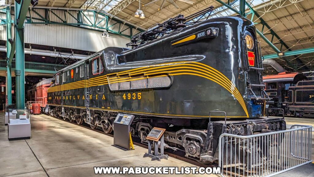 The image captures a majestic Pennsylvania Railroad locomotive, model GG1 4935, displayed prominently at the Railroad Museum of Pennsylvania in Strasburg, Lancaster County. This classic electric locomotive, painted in black with bold yellow striping, reflects the distinctive design and engineering prowess of the mid-20th century. It features a streamlined body with rounded ends and large driver's windows, characteristic of the GG1 series known for their speed and power. The locomotive is shown within the museum's spacious exhibit hall, under a high industrial ceiling, where it is surrounded by other historic rail artifacts. This powerful representation of American railroading heritage stands as a centerpiece in the museum, highlighting the technological advancements and aesthetic considerations of its era.