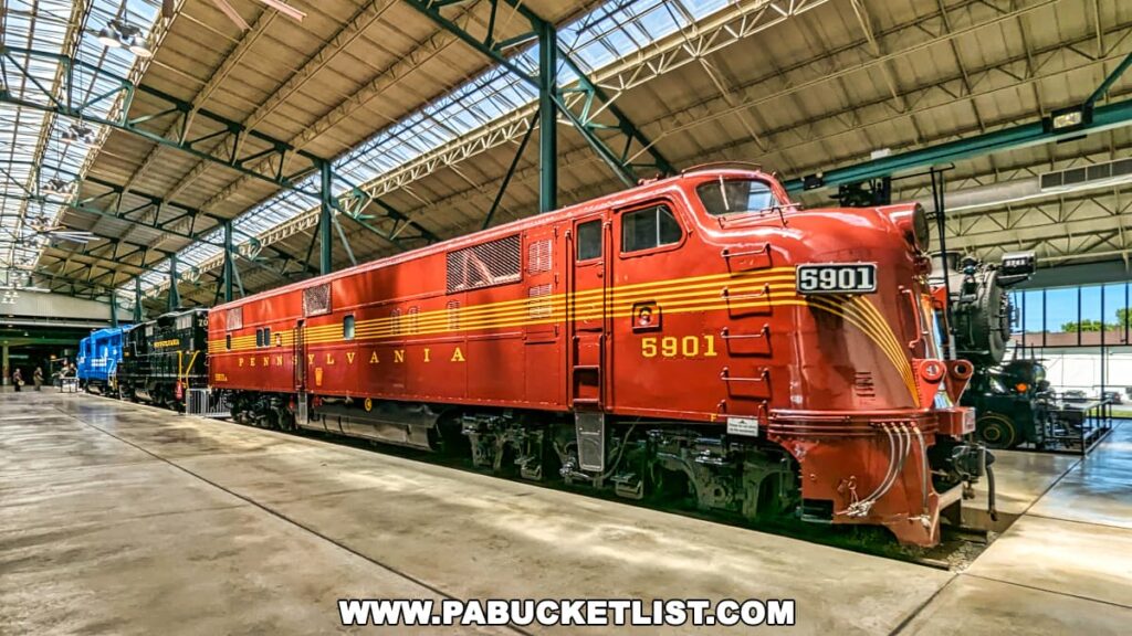 The image captures a stunning red Pennsylvania Railroad locomotive, model 5901, on display at the Railroad Museum of Pennsylvania in Strasburg, Lancaster County. This diesel-electric locomotive, with its vibrant red body and striking yellow striping, exemplifies the classic design of mid-20th-century American rail engineering. The locomotive features sleek, rounded lines and a distinctive front nose, characteristic of streamlined design from that era. It is displayed indoors within a vast exhibit hall, illuminated by natural light streaming through the large ceiling skylights, and is part of a larger collection of historic trains, reflecting the rich railroad heritage preserved at the museum.