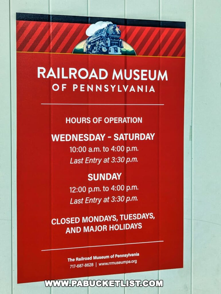 The image features a vibrant red information sign for the Railroad Museum of Pennsylvania located in Strasburg, Lancaster County. The sign includes the museum’s logo with a graphic of a steam locomotive at the top and provides details about the hours of operation. It lists the museum as open Wednesday through Saturday from 10:00 a.m. to 4:00 p.m., with last entry at 3:30 p.m., and on Sundays from 12:00 p.m. to 4:00 p.m., with the same last entry time. It also notes that the museum is closed on Mondays, Tuesdays, and major holidays. The sign is clear, well-organized, and designed to be easily readable, providing essential visitor information in a concise format.