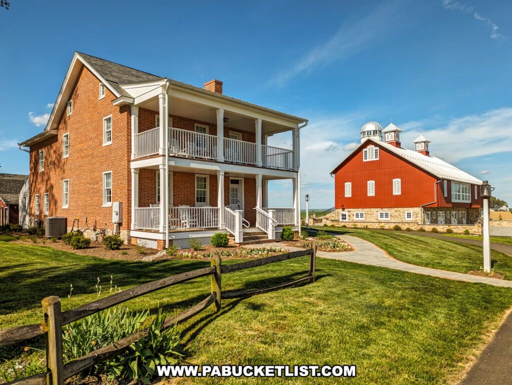 A picturesque farmhouse and barn at the Red Caboose Motel in Lancaster County, PA. The two-story farmhouse is constructed of red brick with white trim, featuring two levels of porches with white railings. The well-manicured lawn and flower beds enhance its charm. In the background, the restored red barn with a metal roof and cupolas, previously described, is visible. A paved walkway connects the two buildings, and a rustic wooden fence adds to the scenic, rural ambiance. The sky is bright blue with a few clouds, highlighting the beautiful weather and setting.