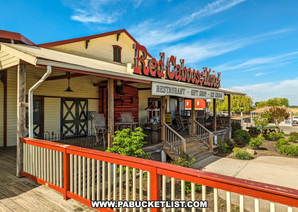 The front entrance of the Red Caboose Motel in Lancaster County, PA, featuring a rustic wooden porch with rocking chairs, and a large sign that reads "Red Caboose Motel." The building has a vintage look with yellow siding, green barn-style doors, and red accents. A sign below the main sign advertises a restaurant, gift shop, and ice cream. The porch is adorned with plants and bushes, and a wooden ramp and stairs provide access. The clear blue sky and well-maintained landscaping enhance the inviting atmosphere of the motel.