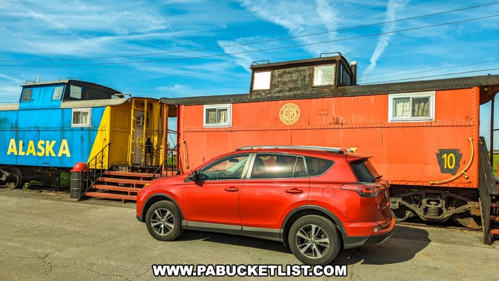 A vibrant red car parked in front of two repurposed cabooses at the Red Caboose Motel in Lancaster County, PA. The caboose on the left is painted blue and yellow with "ALASKA" written on the side, while the caboose on the right is orange with "SOUTHERN" and the number 10. Both cabooses have stairs leading up to their entrances. The sky is clear with streaks of clouds, and the scene is brightly lit by the sun, showcasing the unique and colorful accommodations offered by the motel.