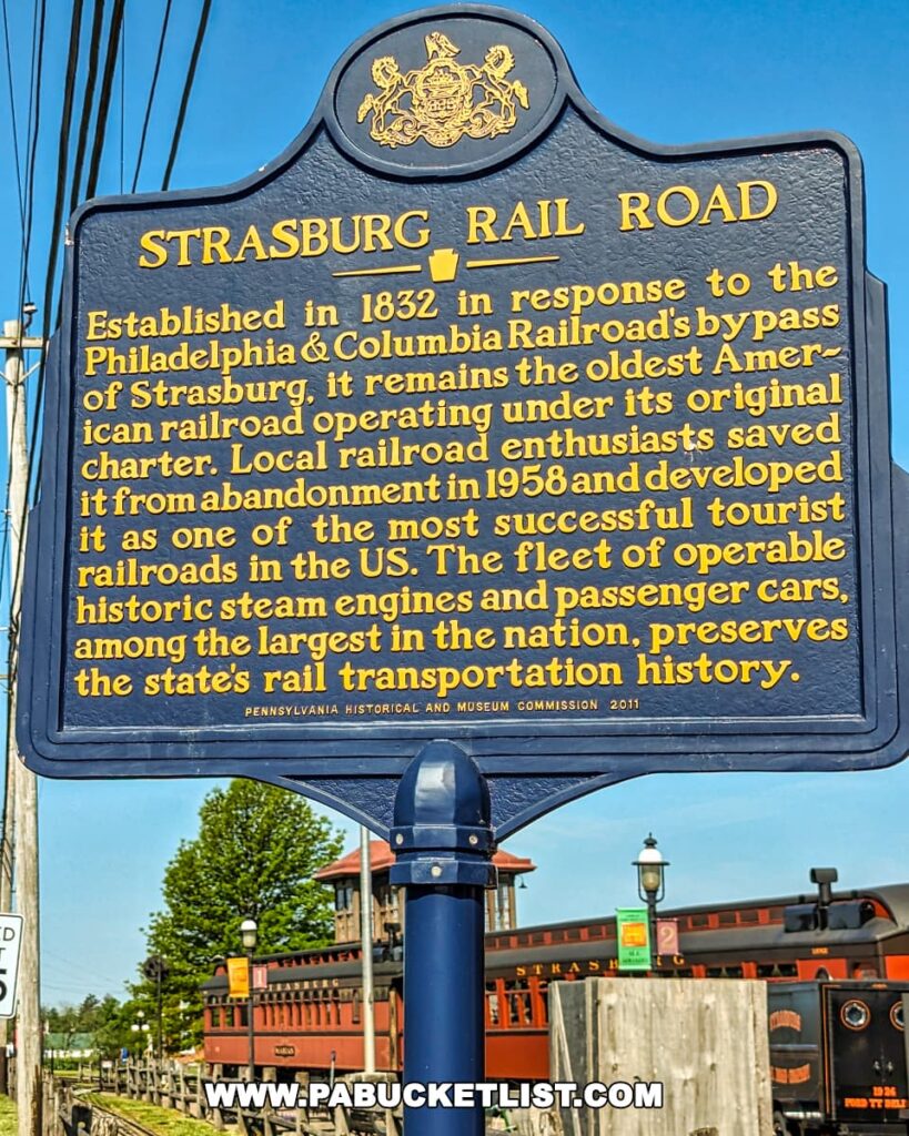 A blue and gold plaque mounted on a metal pole in front of a red and black steam locomotive. The plaque commemorates the Strasburg Railroad, the oldest operating railroad under its original charter in the United States. Text on the plaque describes the history of the railroad and its importance to the state’s rail transportation history.