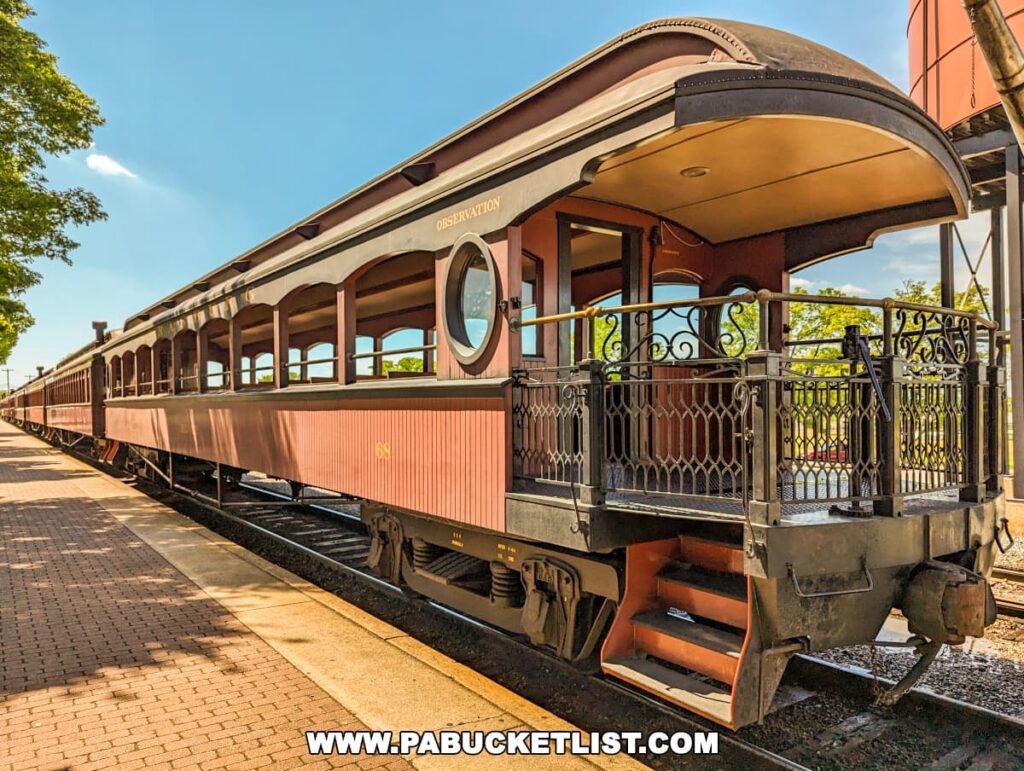 Elegant observation car at the Strasburg Railroad, Lancaster PA, featuring an open rear platform with ornate wrought iron railing and a rounded roof. The car is painted in a deep red with gold trim and large rounded windows, designed to offer passengers expansive views of the surrounding countryside. The railway track and station environment are visible in the background, emphasizing the historic and charming setting of this heritage railroad.