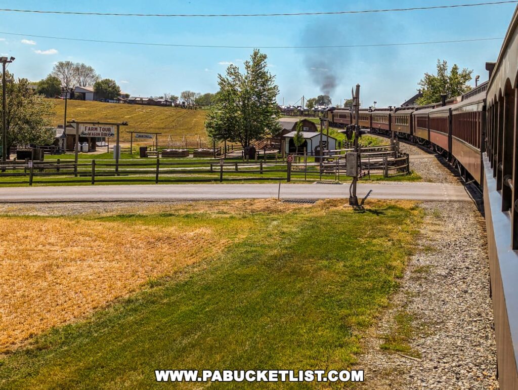 View from the side of a moving train at Strasburg Railroad, overlooking a scenic route in Lancaster County, PA. The landscape features a green grassy area alongside a dried field, with a sign labeled "Farm Tour at Cherry Crest Adventure Farm" visible across the road. In the distance, a smoke plume rises from another train amidst a backdrop of rolling hills and farm structures, capturing the essence of rural Pennsylvania. The train cars curve gently, leading the eye through the tranquil pastoral setting.