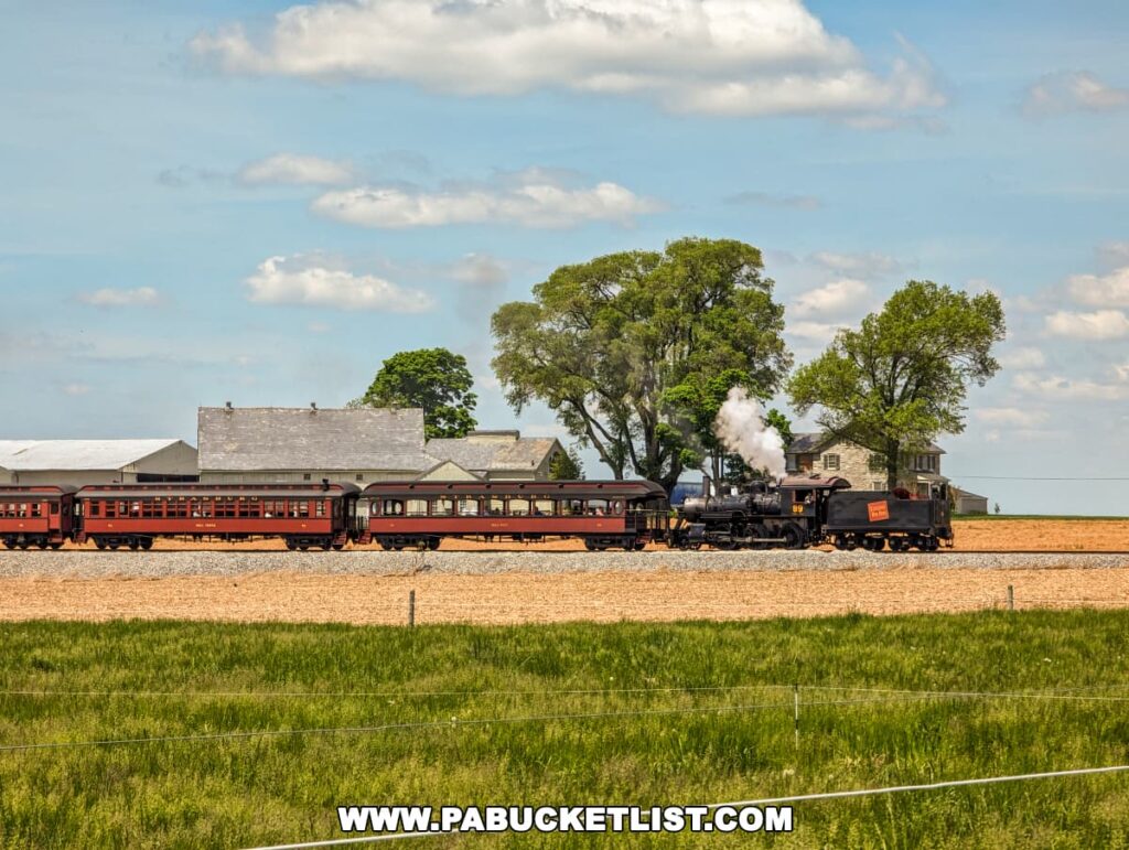 A classic steam locomotive, marked with the number 89, pulls a series of red passenger cars across the rural landscape of Lancaster County, PA. The train is depicted steaming along the tracks with a plume of white smoke, surrounded by vibrant green fields under a clear blue sky. In the background, traditional farm buildings and a farmhouse add a rustic touch to the picturesque scene at Strasburg Railroad.