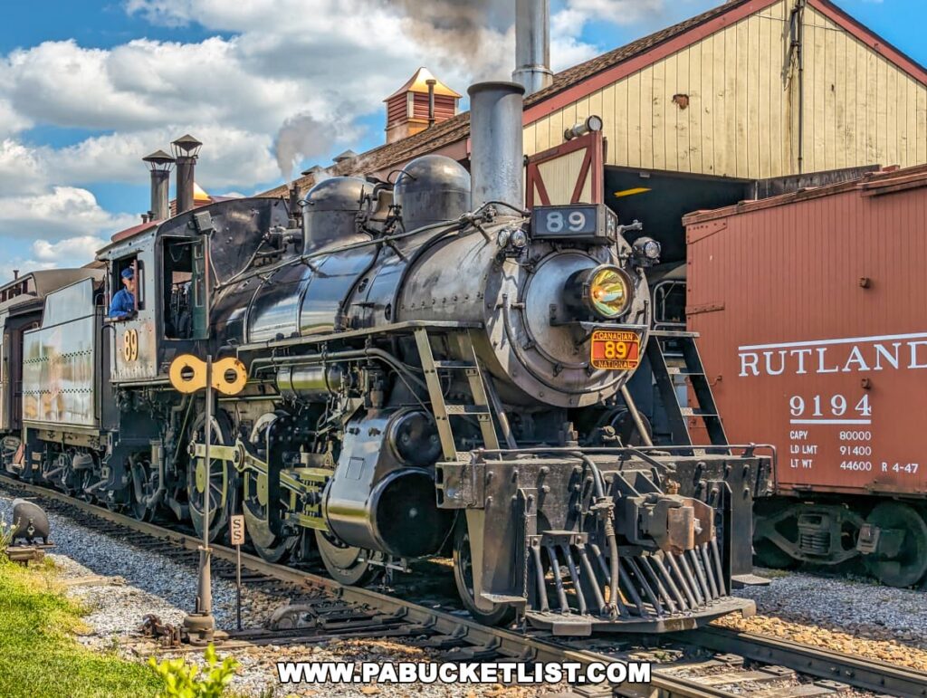 A vintage steam locomotive, numbered 89, is displayed at Strasburg Railroad in Lancaster, PA. The train, marked as "Canadian National 89," features a black and silver exterior and is actively steaming, indicating it is operational. The engineer is visible in the cab, wearing a blue shirt. This scene is set against the backdrop of an old yellow train depot with a sign reading "Rutland 9194" on a nearby red freight car, highlighting the historical atmosphere of the railroad.
