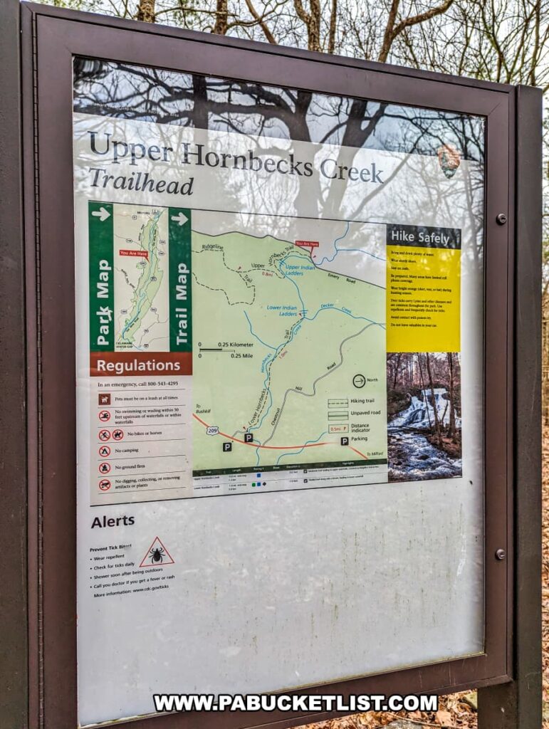 A photo of the trailhead sign for Upper Hornbecks Creek in Pike County, Pennsylvania. The sign features a detailed trail map showing the route to Upper Indian Ladders Falls, along with safety tips, regulations, and alerts. The surrounding area includes bare branches and hints of forest, indicating a natural and wooded environment. The information board provides hikers with essential details for safely exploring the trails and reaching the waterfalls along Upper Hornbecks Creek.