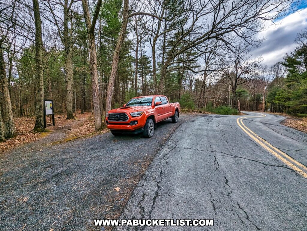 A view of the parking area at the trailhead for Upper Indian Ladders Falls in Pike County, Pennsylvania. A red truck is parked on the side of a winding road, with a forested area in the background. The trailhead sign is visible near the trees, marking the entrance to the hiking path leading to the waterfall. The surrounding forest features a mix of evergreen and deciduous trees, creating a natural and inviting atmosphere for hikers.