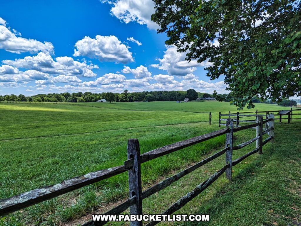 A scenic view of a vast, green meadow at Brandywine Battlefield Park in Chester County, Pennsylvania, with a rustic wooden fence running along the right side. The sky is bright blue with fluffy white clouds, and a few historical buildings are visible in the distance. This location is significant as the site of the Battle of the Brandywine, the largest and longest single-day land battle of the American Revolution.