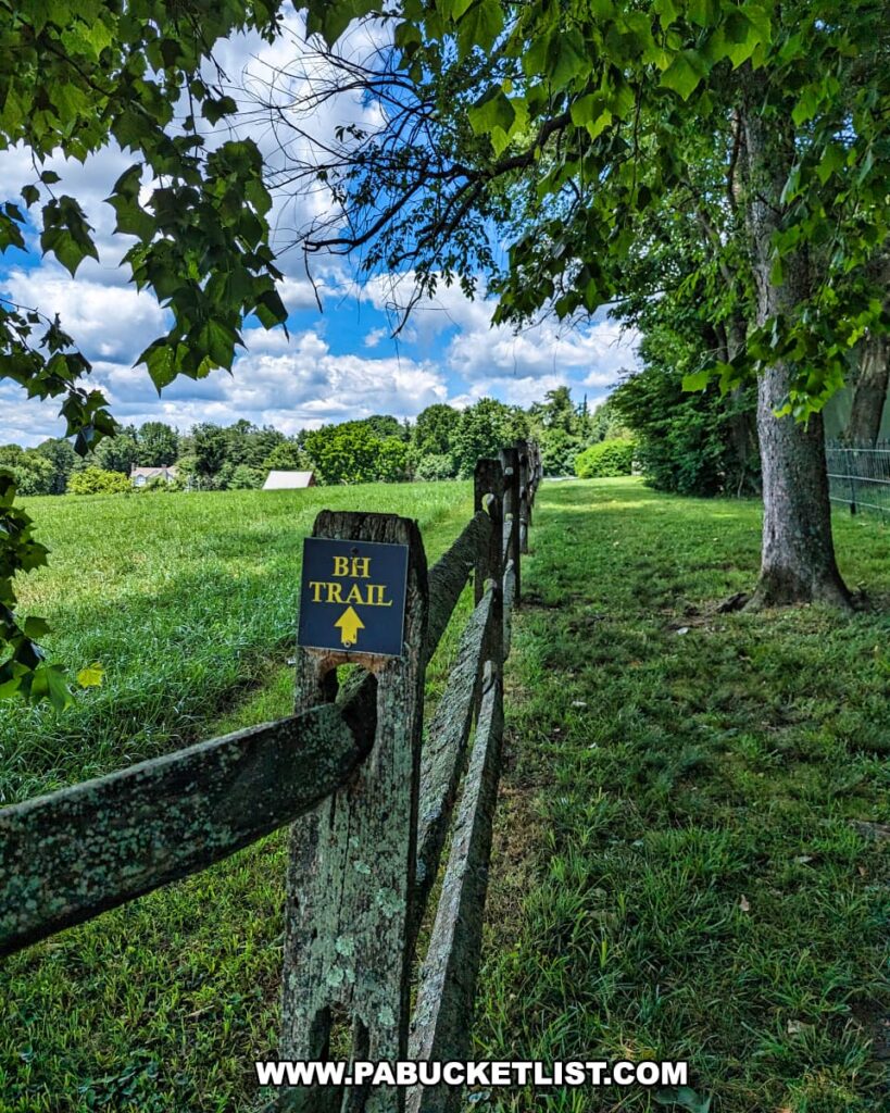 A scenic view along the Brandywine Hill Trail at Brandywine Battlefield Park in Chester County, Pennsylvania. The trail is marked by a rustic wooden fence with a blue and yellow sign indicating "BH Trail" and an arrow pointing forward. The path is shaded by large trees with green leaves, and the trail runs through a grassy field under a bright blue sky with scattered clouds. This serene setting provides a peaceful contrast to the historical significance of the park, where the largest and longest single-day land battle of the American Revolution took place.