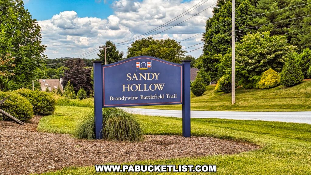 A sign marking the entrance to Sandy Hollow at Brandywine Battlefield Park in Chester County, Pennsylvania, stands prominently beside a road. The sign, painted in dark blue with gold lettering, reads "Sandy Hollow Brandywine Battlefield Trail" and features a historical flag at the top. The surrounding area is well-manicured, with neatly trimmed bushes, grass, and trees, creating a welcoming and serene atmosphere. In the background, residential homes and power lines are visible, set against a partly cloudy sky, highlighting the park's blend of historical significance and modern-day community.