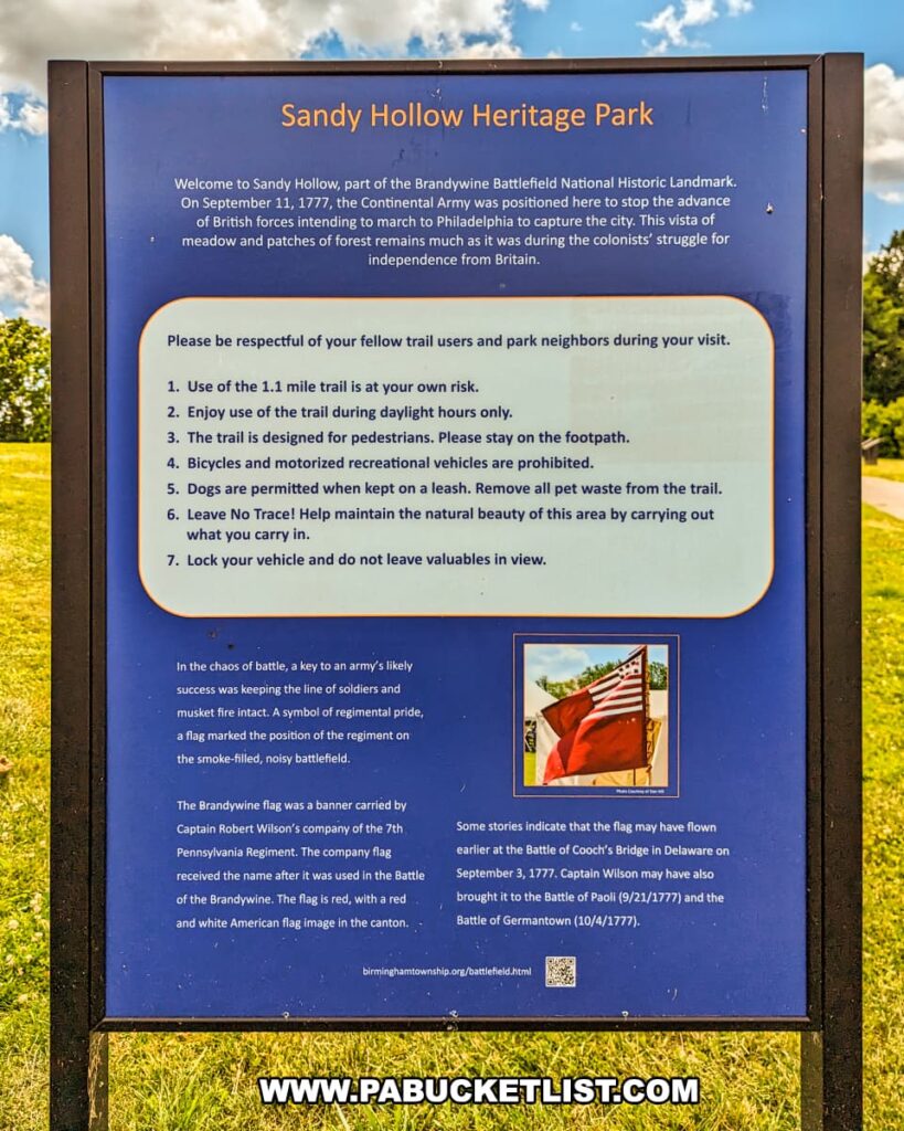 An informational sign at Sandy Hollow Heritage Park, part of Brandywine Battlefield Park in Chester County, Pennsylvania, provides historical context and visitor guidelines. The sign details that on September 11, 1777, the Continental Army was positioned here to stop the British advance during the Battle of the Brandywine. It includes rules for the 1.1-mile trail, such as using the trail during daylight, staying on the footpath, and keeping dogs on a leash. The sign also describes the historical significance of the Brandywine flag, used by Captain Robert Wilson's company, and its role in the battle. The background features a clear blue sky and green grass, enhancing the sign's visibility and appeal.