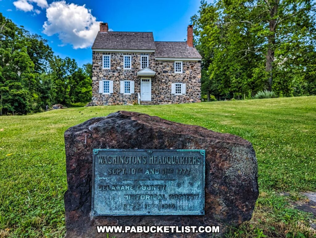 A historic stone house at Brandywine Battlefield Park in Chester County, Pennsylvania, is identified as Washington's Headquarters from September 10th and 11th, 1777. In the foreground, a large rock bears a plaque commemorating this fact, presented by the Delaware County Historical Society on September 16th, 1946. The house stands amid a well-maintained grassy area, surrounded by trees, with a bright blue sky and fluffy white clouds overhead, highlighting its historical significance and picturesque setting.