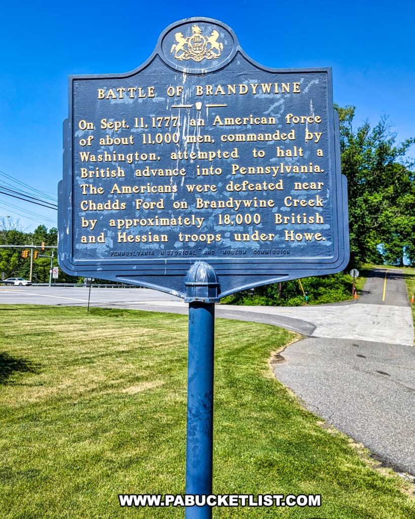 Historical marker at Brandywine Battlefield Park in Chester County, Pennsylvania, commemorating the Battle of the Brandywine on September 11, 1777. The marker details the engagement where an American force of about 11,000 men, commanded by Washington, attempted to halt a British advance into Pennsylvania but were defeated near Chadds Ford by approximately 18,000 British and Hessian troops under Howe. The marker stands along a road with a clear blue sky in the background.
